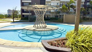 Una piscina con una estructura decorativa central, rodeada de un área ajardinada con plantas y un edificio de varios pisos al fondo.
