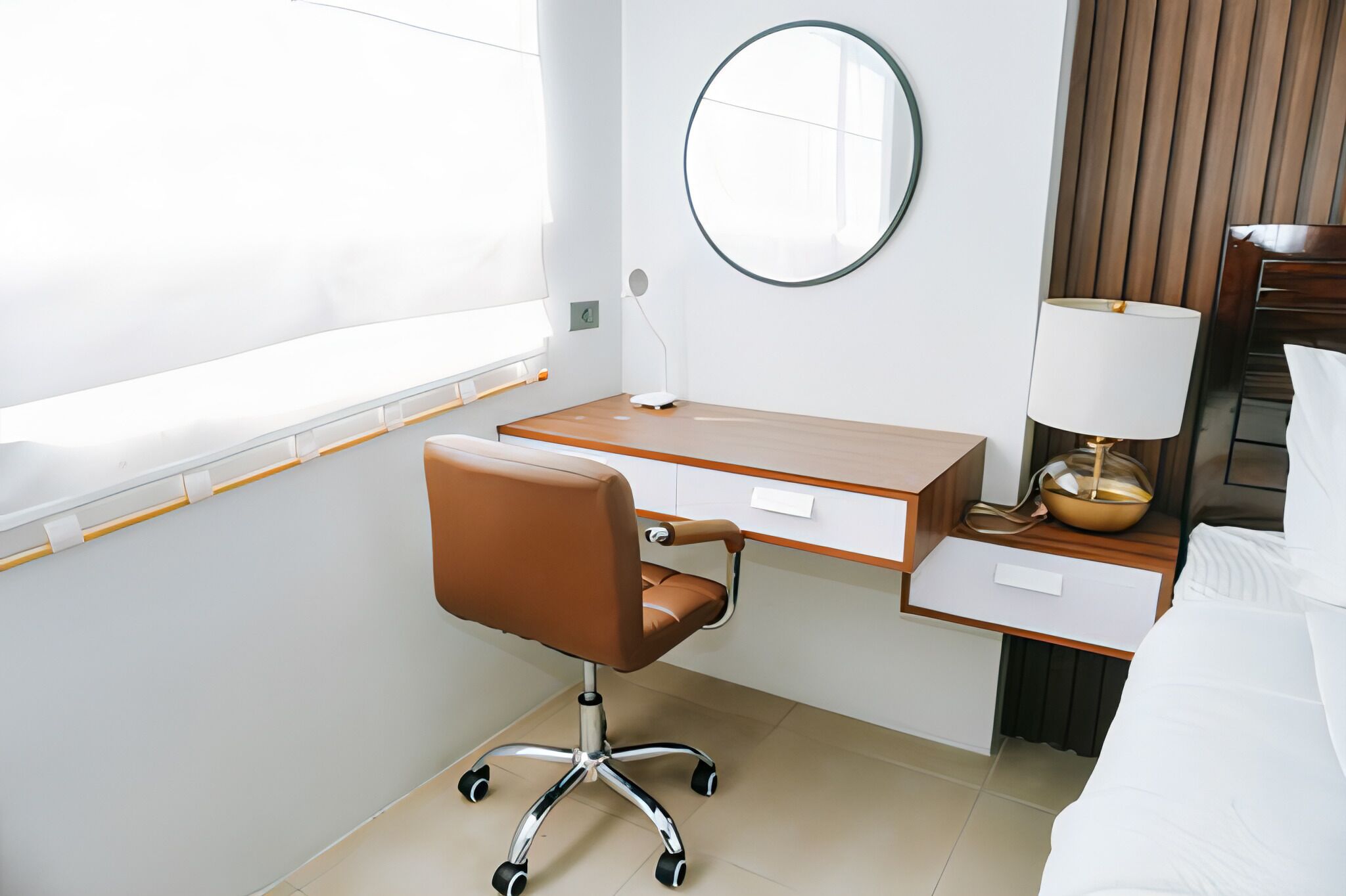 A hotel room with a wooden desk, a brown office chair, a round mirror, a bedside table with a lamp, and a bed with white bedding.