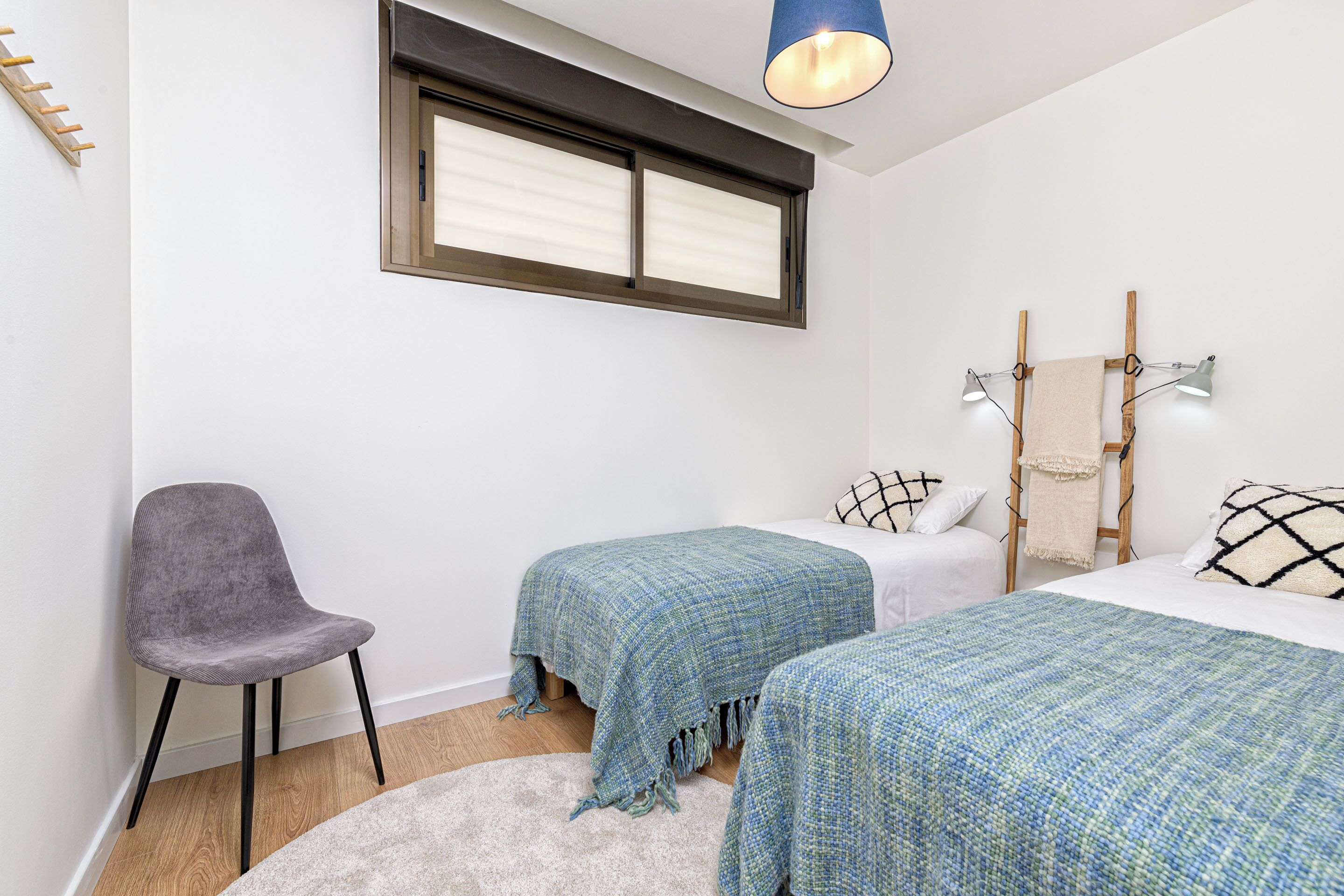 A bedroom with two beds, a chair, a window, and a hanging light fixture.