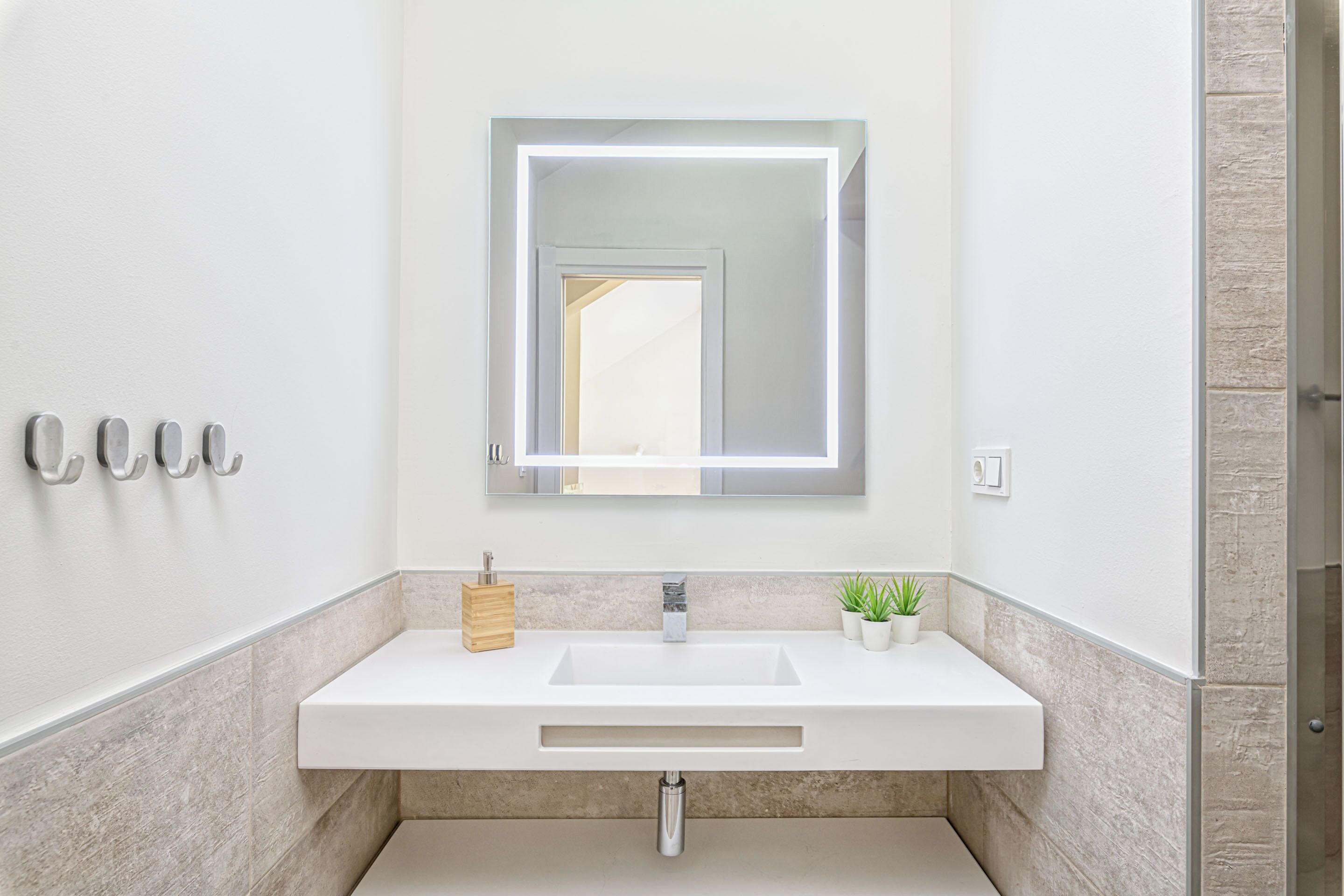 A modern bathroom with a white vanity, a mirror with integrated lighting, a small potted plant, and a wooden soap dispenser.