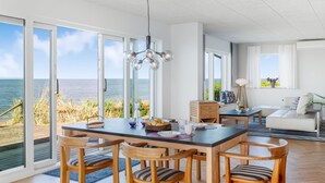 Ett modernt matområde med utsikt över havet, trästolar, ett stort bord i mitten och en hängande lampa.