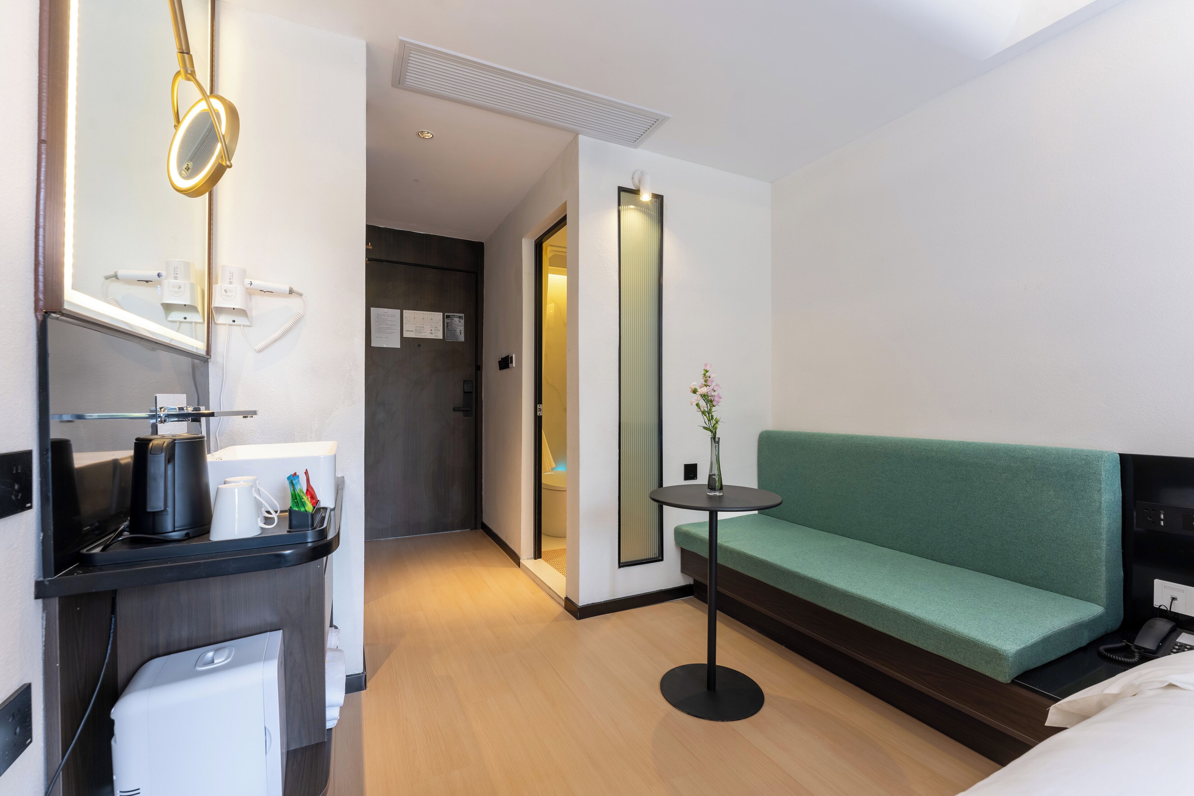 A modern hotel room with a green sofa, a small round table, a desk with a coffee maker, and a mirror.