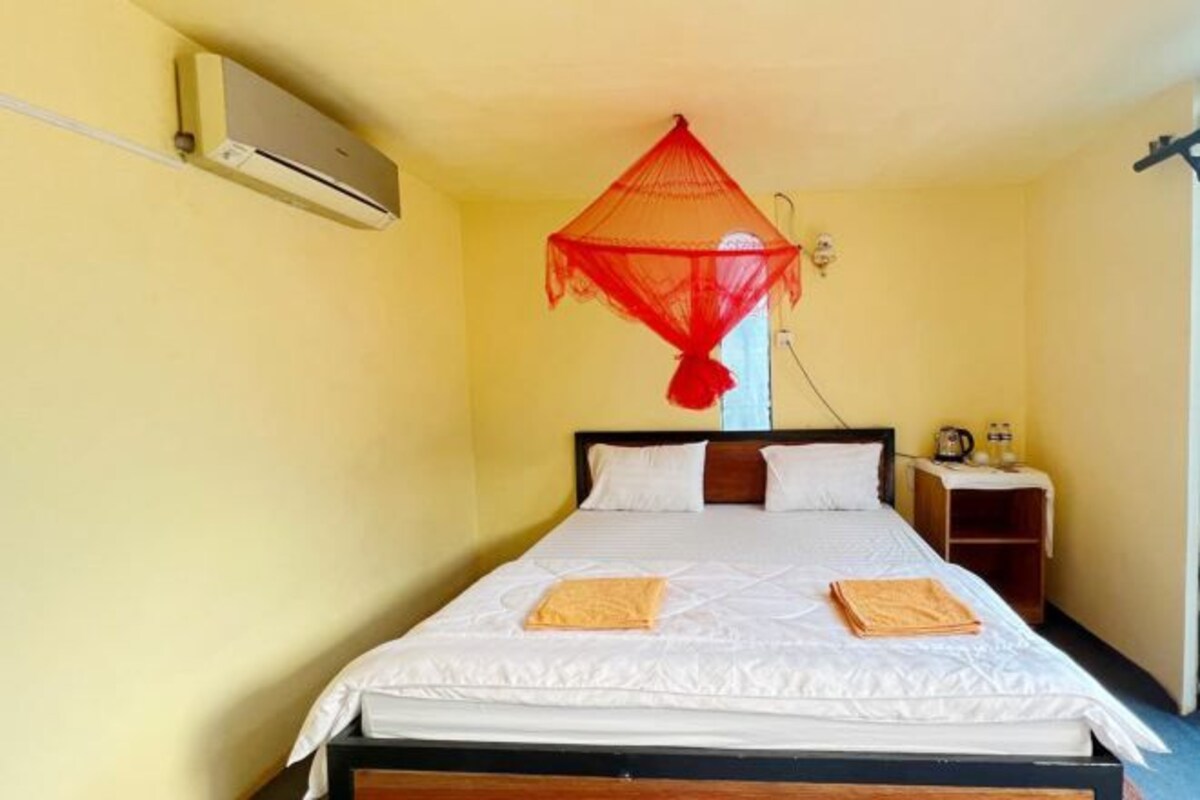 A single bed with white bedding, a red mosquito net, and a small bedside table with a lamp.
