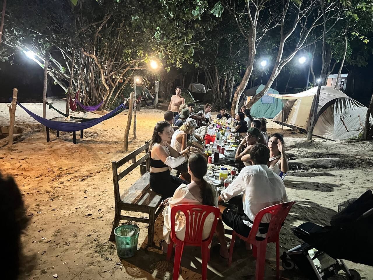 Un groupe de personnes dînant en plein air, la nuit, sous une voûte d’arbres, avec un hamac et des tentes en arrière-plan.