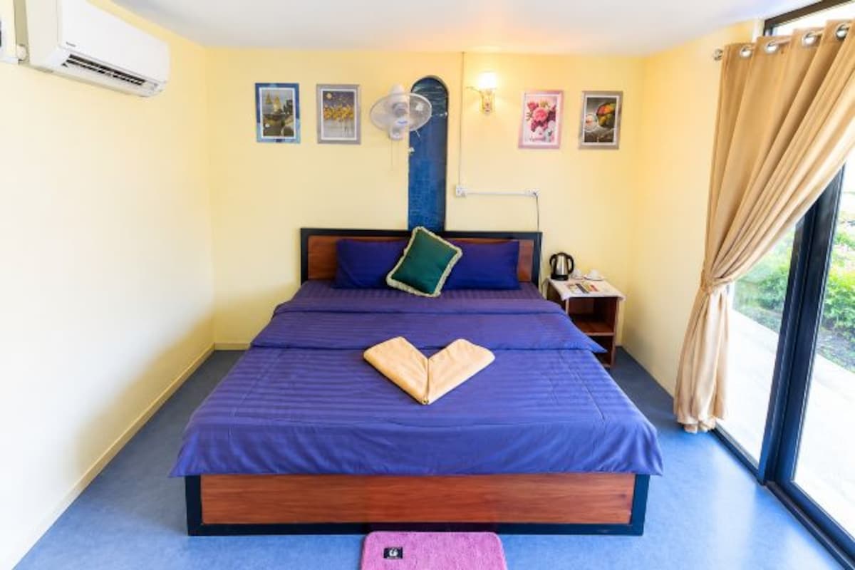 A bedroom with a large bed, a bedside table, a fan, and a window with curtains.