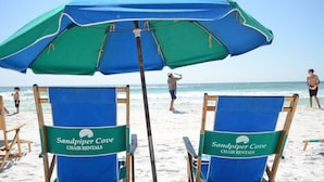 Duas cadeiras de praia com a marca 'Sandpiper Cove Chair Rentals', um grande guarda-sol azul e verde, e um cenário de praia com pessoas ao fundo.