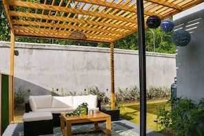 An outdoor patio with a wooden pergola, a white sofa, a wooden table, and decorative hanging lights.