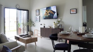A modern living room with a dining area, a television mounted on the wall, and a large window providing natural light.