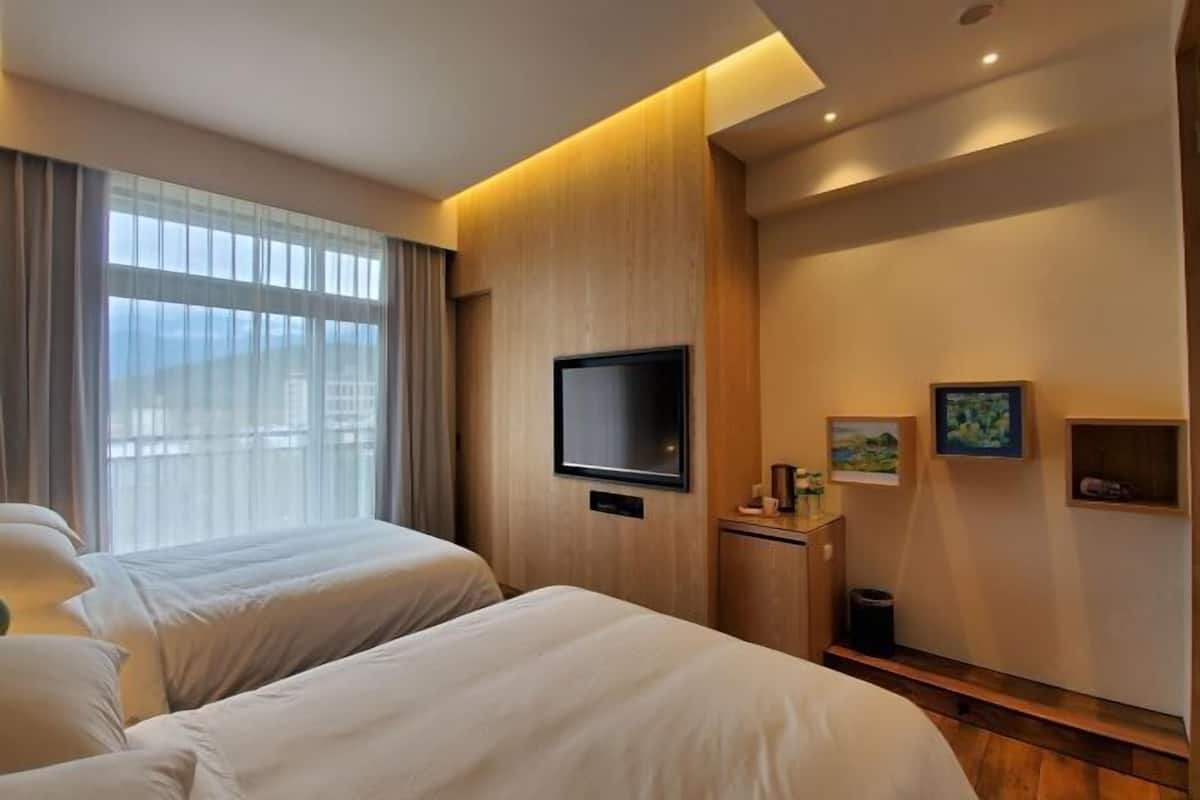 A hotel room with two beds, a TV, a small desk, and a window with curtains.