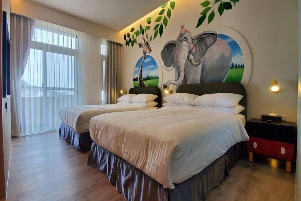 A hotel room with two beds, a large window with curtains, a wall mural of an elephant, and a nightstand with a lamp.