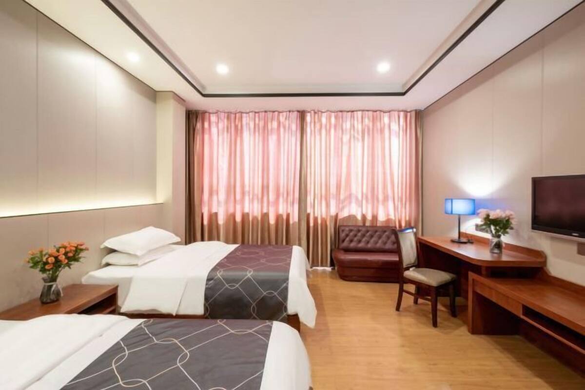A hotel room with two beds, a desk, a chair, a TV, and a large window with curtains.