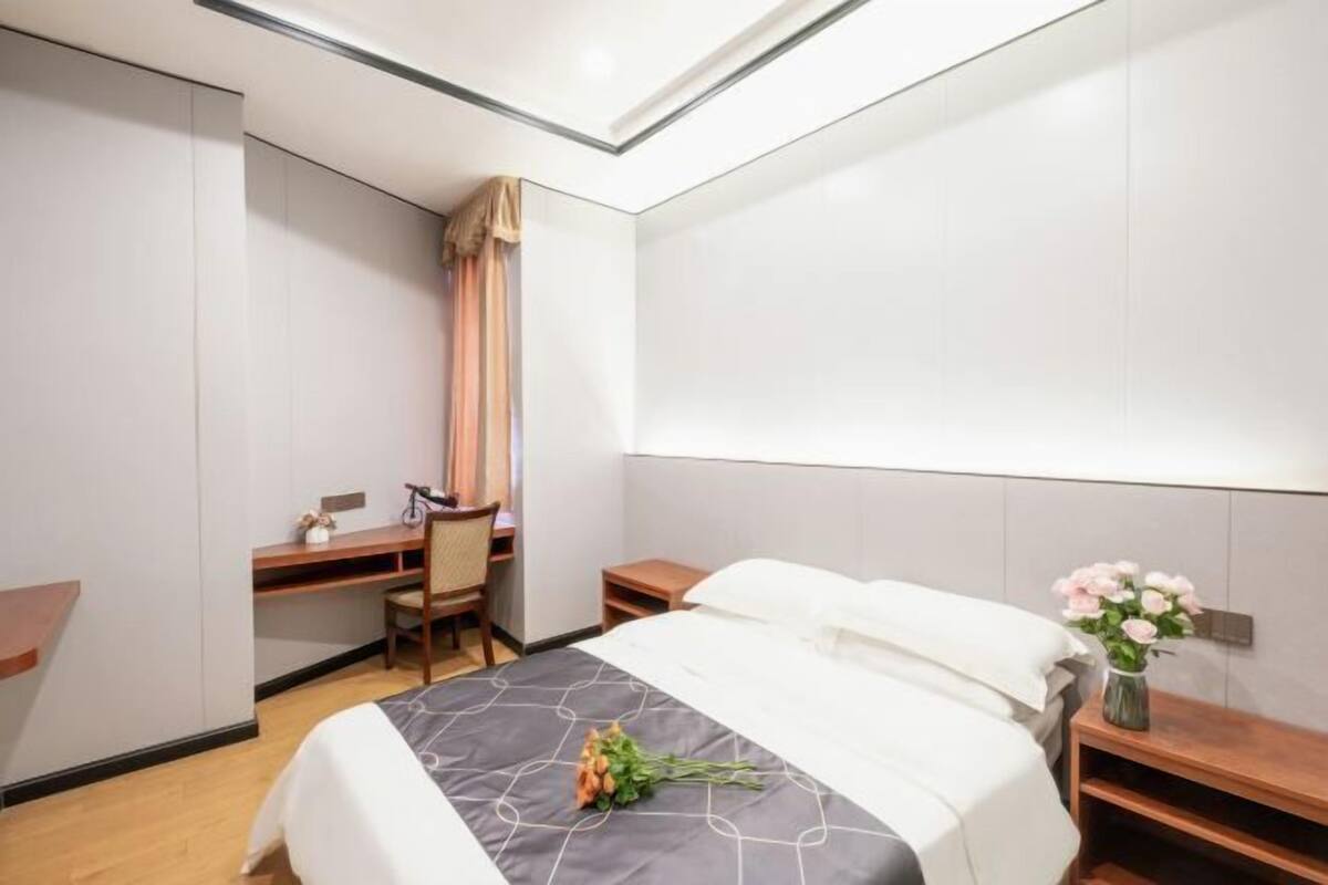 A hotel room with a bed, desk, chair, and a small bedside table with a vase of flowers.