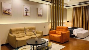 A modern living room with a beige sofa, an orange armchair, and a small round table. There are three framed artworks on the wall and a large window with curtains. - Jiangmen Mintai Hotel (Jiangmen)