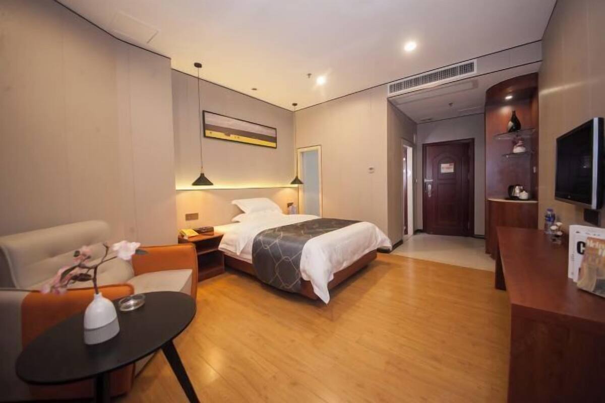 A hotel room with a bed, a desk, a chair, a small table with a vase, and a television.
