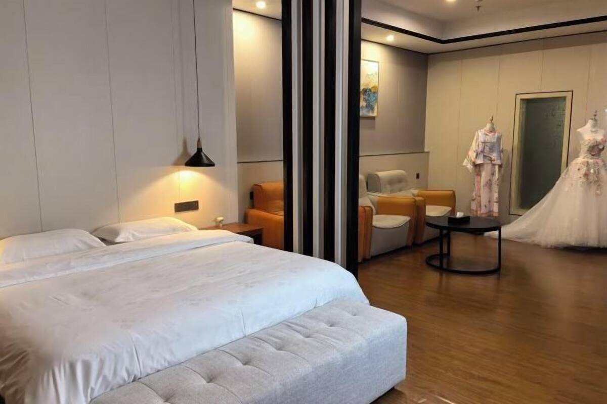 A modern hotel room with a large bed, a seating area with chairs, a small table, and a mannequin in a wedding dress.