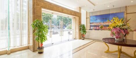 Un hall d’hôtel avec une entrée dotée d’une porte en verre, des plantes en pot, un comptoir de réception et un grand tableau accroché au mur.