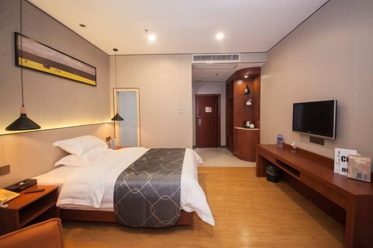 A hotel room with a bed, bedside table, lamp, TV, and a wooden bench.