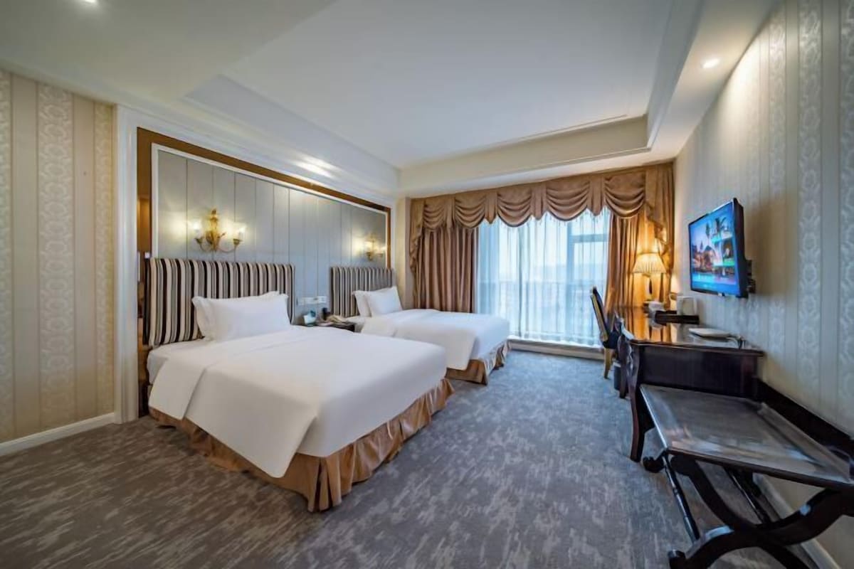 A hotel room with two beds, a desk, a chair, a television, and a large window with curtains.