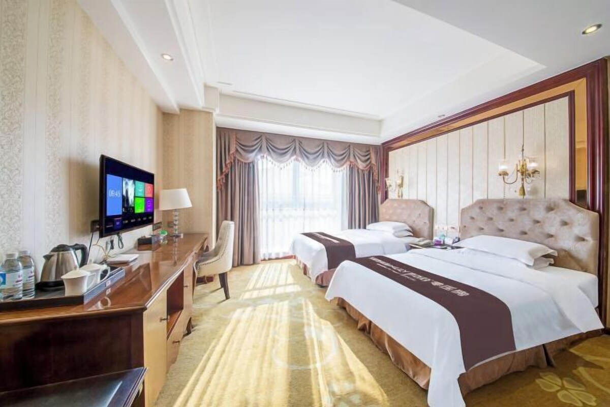 A hotel room with two beds, a desk with a television, a chair, and a large window with curtains.