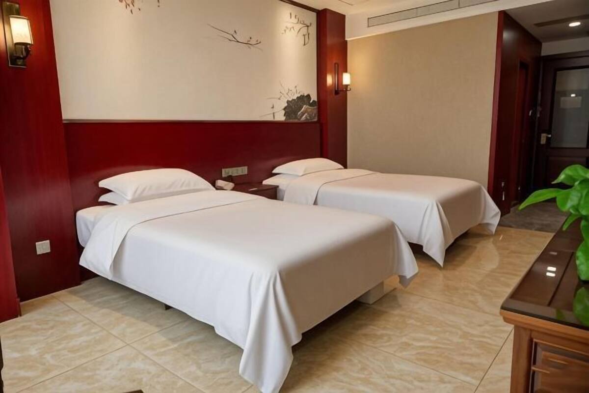 A hotel room with two beds, a nightstand, a wall-mounted light, and a plant.