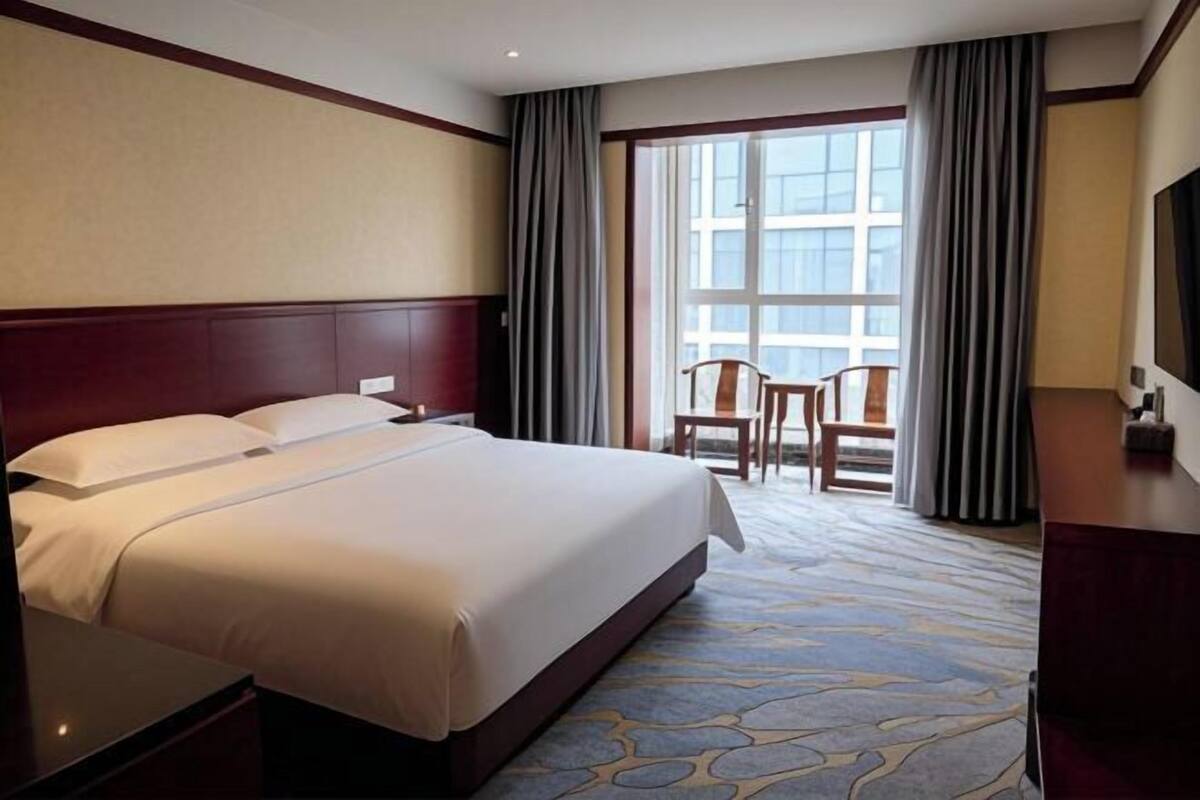 A hotel room with a large bed, a desk, a chair, and a view of the outside through a window.