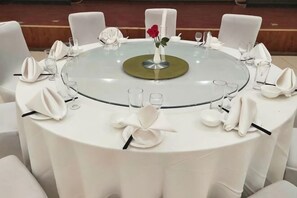 Une table ronde recouverte d’une nappe blanche, dressée pour un repas, entourée de chaises blanches.