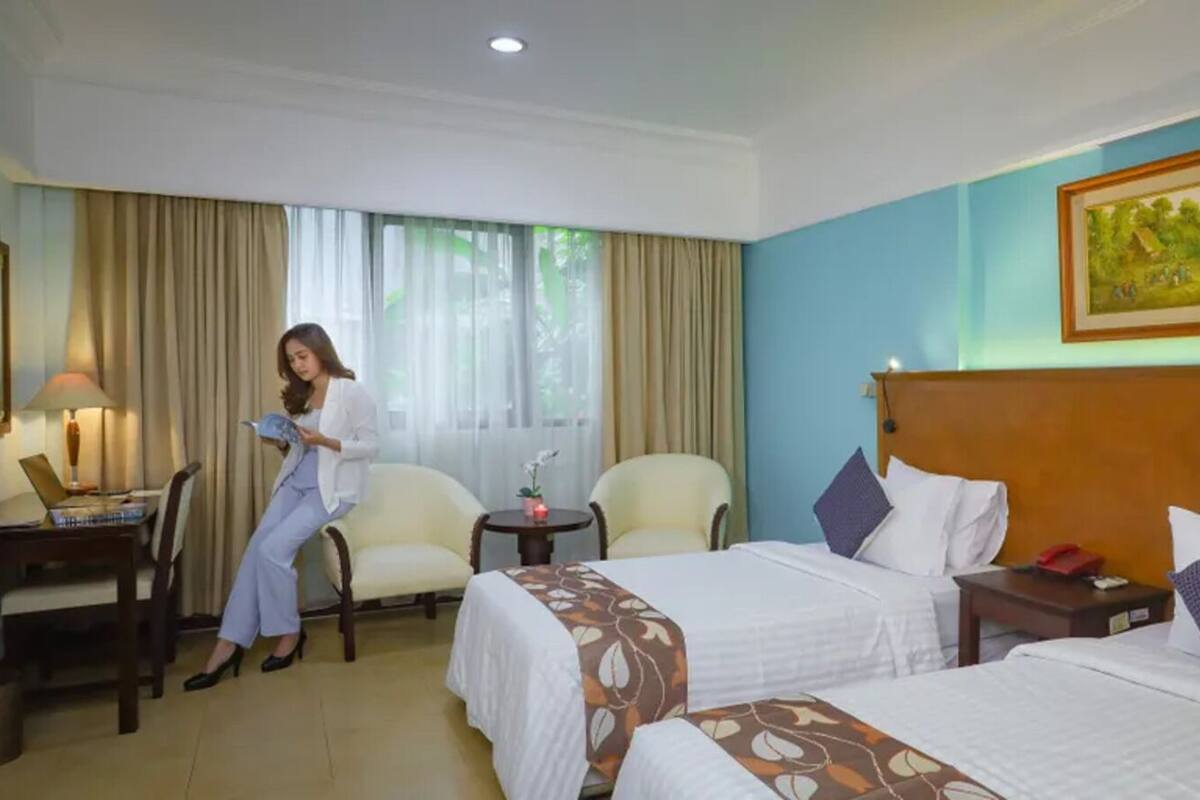 A hotel room with two beds, a desk, a chair, and a small table with a lamp.