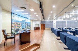 Un salón de conferencias con una pantalla grande mostrando un paisaje urbano, una mesa de recepción y filas de mesas con manteles azules.