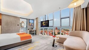 A hotel room with a bed, a sofa, a small table, a TV, and a large window with a city view.