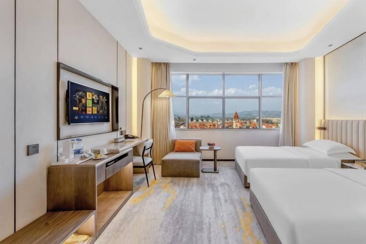 Habitación de hotel con escritorio, dos camas, televisor, vista a la ciudad y un ventanal grande.