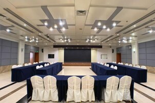 Un salón de conferencias con mesas largas, sillas, un escenario y una pantalla para proyector.