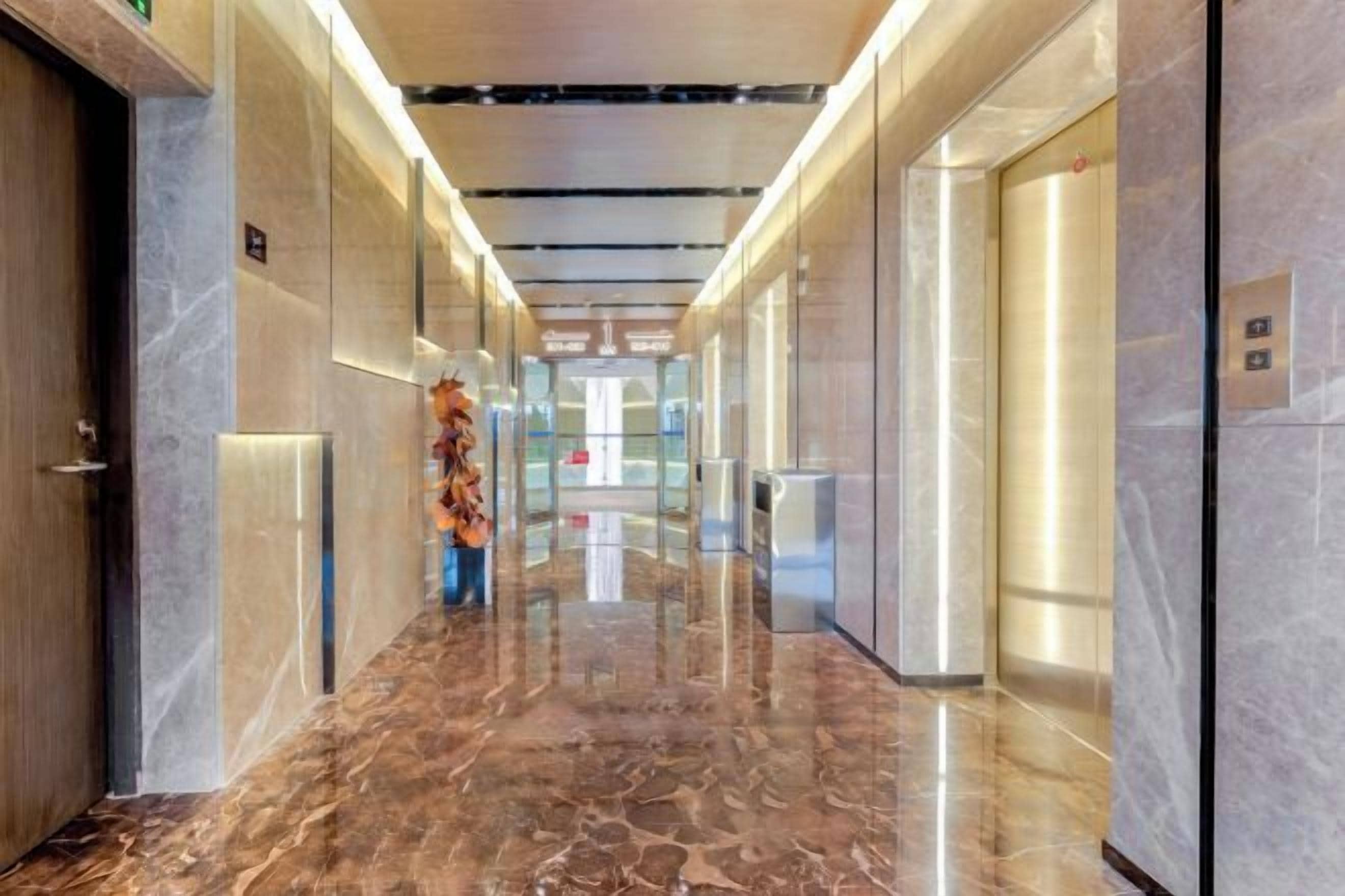 Un pasillo moderno de hotel con suelo de mármol, puertas de vidrio y un ascensor central.