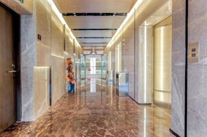 Un pasillo moderno de hotel con suelo de mármol, puertas de vidrio y un ascensor central.