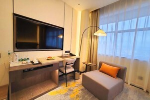Habitación de hotel con cama, escritorio con computadora, televisión, lámpara y ventana con cortinas.