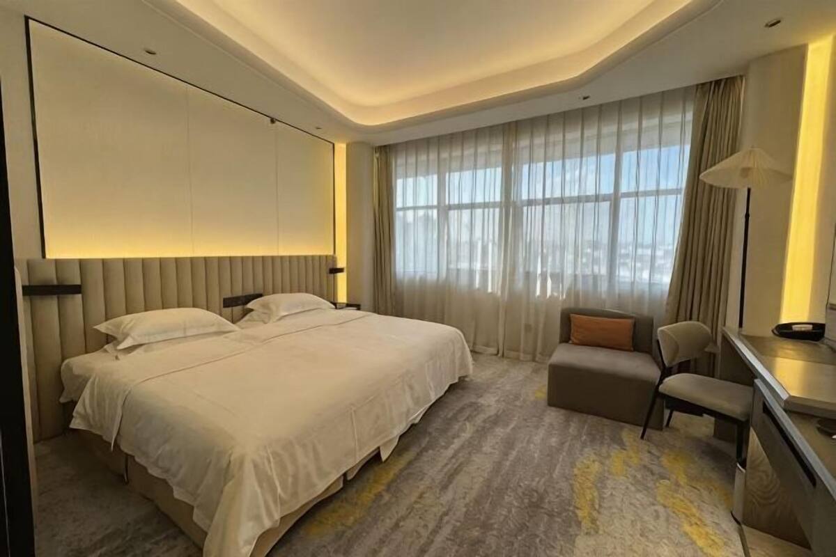 Habitación de hotel con una cama grande, un escritorio, una silla y vistas de la ciudad a través de amplias ventanas.