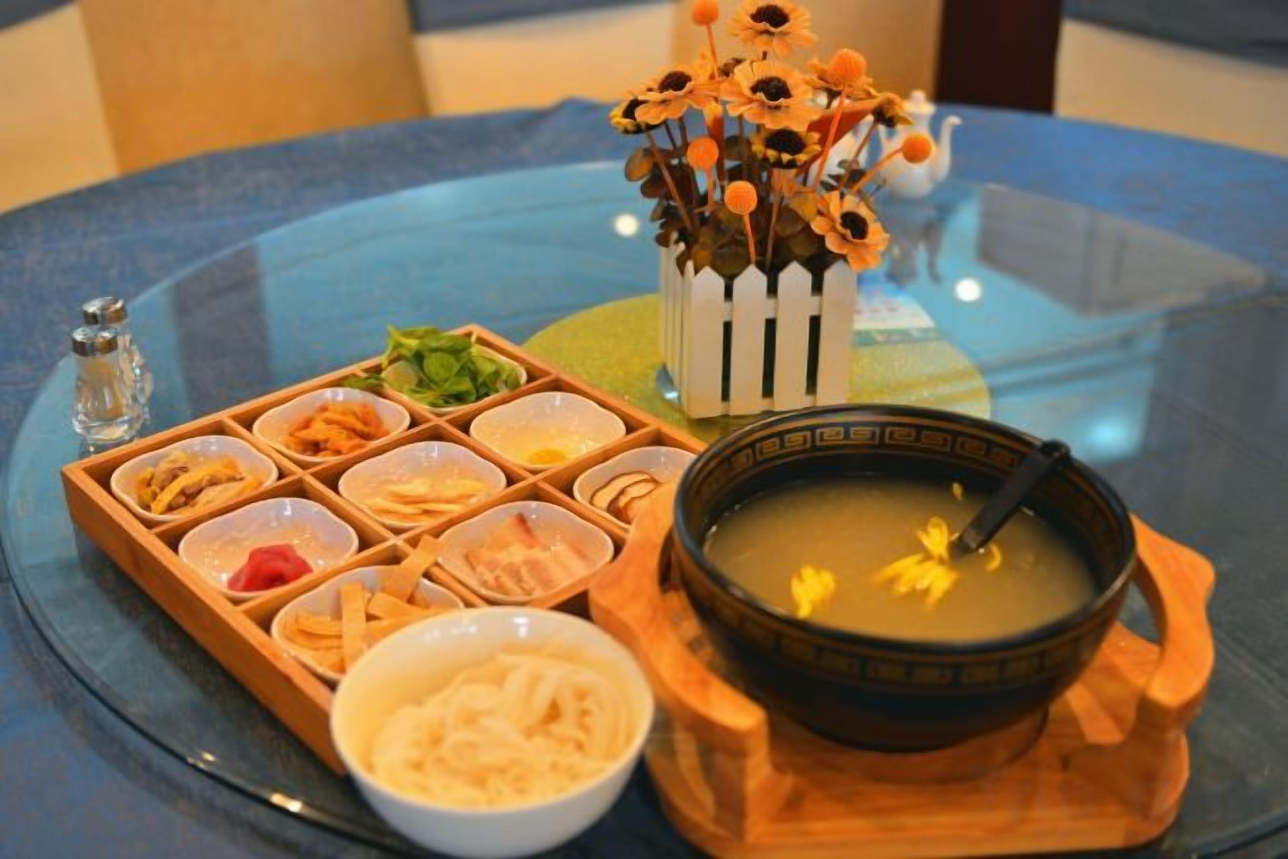 Una mesa con un tazón de sopa, una bandeja de alimentos y un florero decorativo.