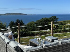A seaside terrace with wicker furniture, a table with wine glasses, and a rope barrier. - Mull Tranquility Retreat (Isle of Mull)