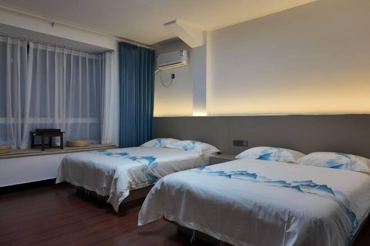 A hotel room with two beds, a window with sheer curtains, an air conditioning unit, and a bedside table.