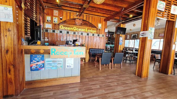 Egy recepciós pult, rajta a „Green Ocean Lodge” felirattal, és egy külön „Recepció” tábla. A helyiségben fából készült székek és asztalok találhatók.