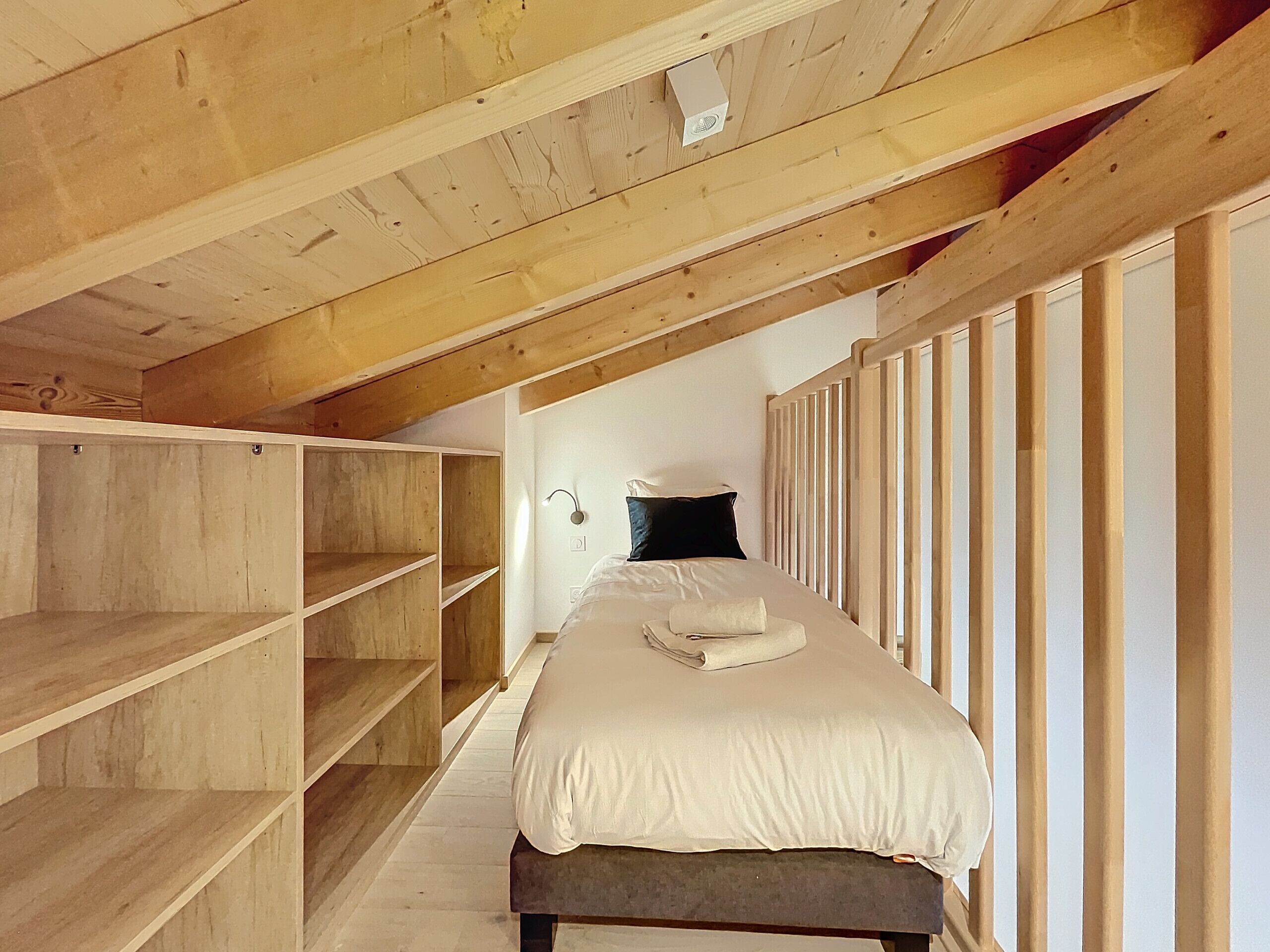 A bedroom with a wooden sloped ceiling, a bed with a white mattress and pillows, and built-in wooden shelving.