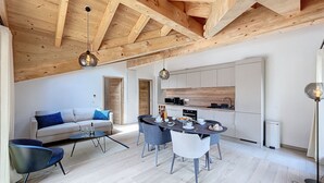 A modern living room with a sofa, dining table, and chairs, a kitchen with wooden cabinets, and a high ceiling with exposed beams. - Cedrela-appartement-5/7pers-Centre station (Les Gets)