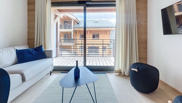 Un salon moderne avec un canapé, une petite table et un pouf noir. Une grande fenêtre offre une vue sur un bâtiment en bois situé à l’extérieur.
