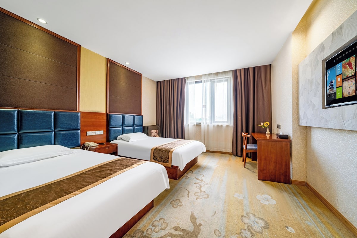 Ein Hotelzimmer mit zwei Betten, einem Schreibtisch, einem Stuhl, einem Fernseher und einem großen Fenster mit Vorhängen.