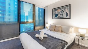 Une chambre d’hôtel moderne dotée d’un grand lit, offrant une vue sur la ville grâce à de larges fenêtres du sol au plafond, et agrémentée d’une peinture abstraite contemporaine au mur.