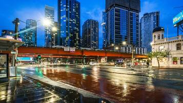 A cityscape with tall buildings, a wet street reflecting lights, and a pedestrian crossing.