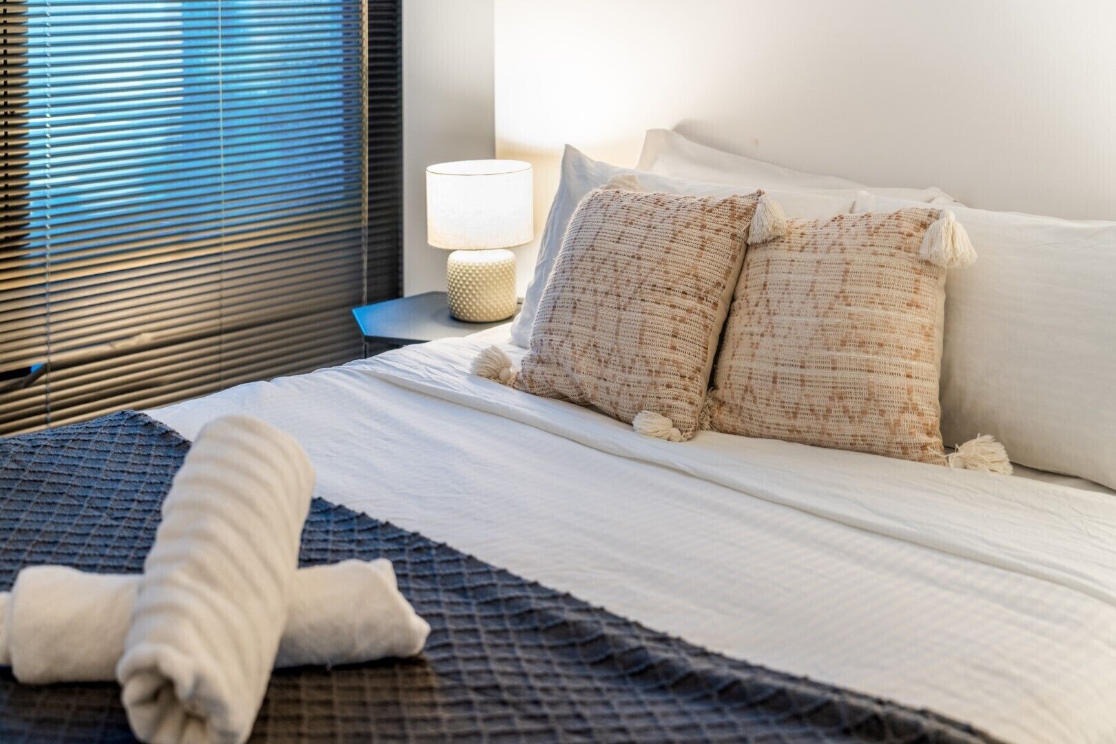 A neatly made bed with a white comforter and two beige pillows, a blue blanket, and a rolled white towel on a textured blue bedspread.