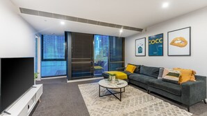 Un salon moderne avec un canapé gris, une table basse et un téléviseur à écran plat. De grandes fenêtres équipées de stores laissent entrer la lumière, et des œuvres d’art ornent les murs.