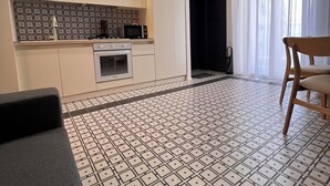 Una cucina moderna con mobili bianchi, un forno a microonde e un forno. C'è un'area pranzo con una sedia di legno e un tavolo. Il pavimento presenta un motivo a piastrelle geometriche.