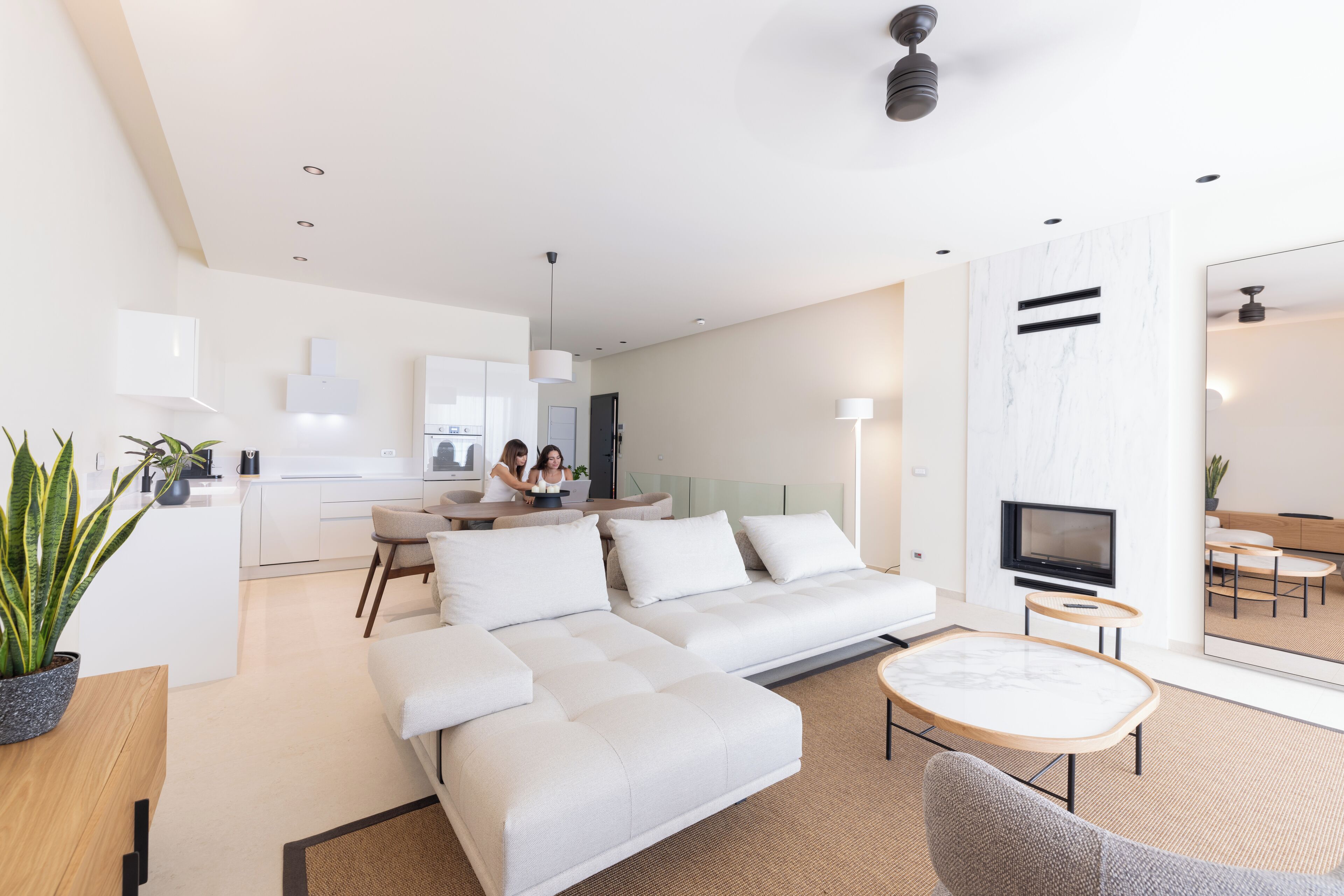 A modern living room with a sofa, coffee table, and TV. There is a kitchen area in the background with a dining table and chairs.