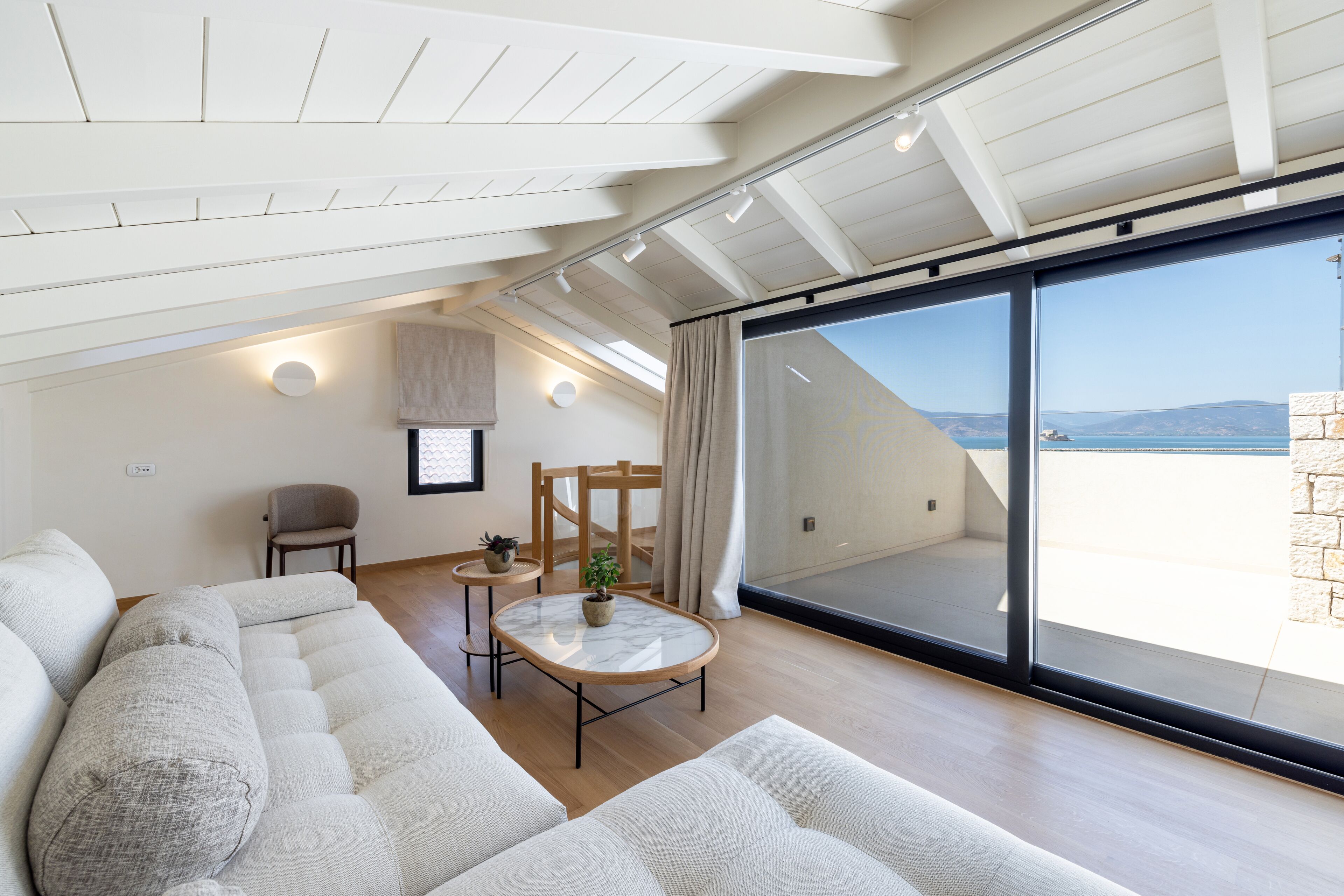 Un dormitorio estilo loft moderno con un gran ventanal corredera que da a un balcón, una zona de estar acogedora con un sofá y una mesita, y una estructura de techo de madera.